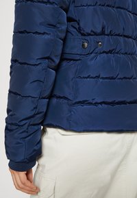 Granatowa kurtka puchowa z poziomym przeszyciem, zapinana na zatrzaski, z prążkowanymi mankietami, noszona na jasnych spodniach.