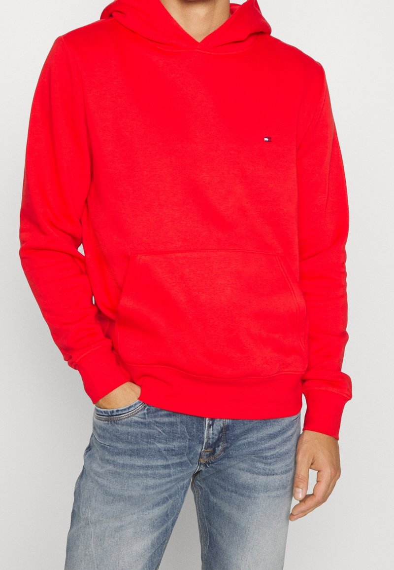 Sweat à capuche rouge avec poche avant porté par une personne en jean bleu délavé, main reposant dans la poche.