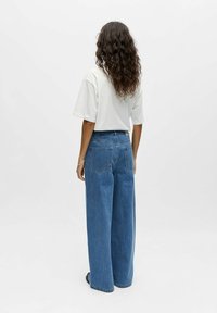 Femme aux longs cheveux bouclés portant un t-shirt blanc à manches courtes et un jean bleu taille haute à jambes larges, debout de dos.