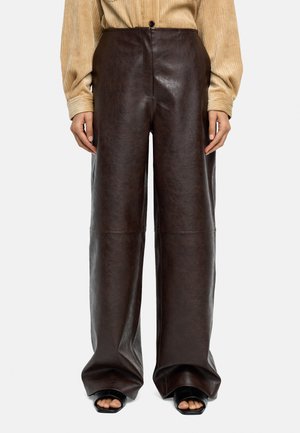 Osoba nosząca wysokie, szerokie spodnie ze skóry w ciemnobrązowym kolorze, połączone z beżowym topem z długim rękawem oraz czarnymi sandałami na kwadratowym obcasie z odkrytymi palcami.