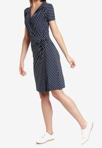 Abito avvolgente a righe navy con maniche corte, caratterizzato da un dettaglio con fiocco in vita. Abbinato a sneakers bianche con lacci.