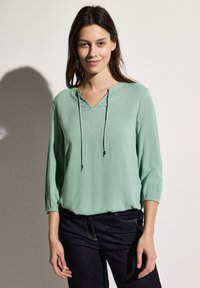 Blouse légère vert menthe avec un col en V, élastiques aux manches et un détail de cordon de serrage. Associée à un jean foncé.