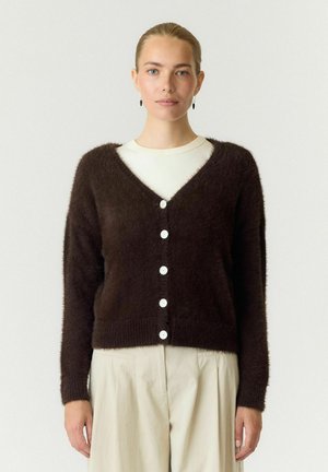 Femme à la peau claire et aux cheveux blonds attachés porte un cardigan marron foncé duveteux boutonné par-dessus un haut blanc et un pantalon beige.