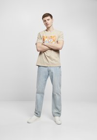 Beige bomull t-skjorte med et fargerikt Bugs Bunny-trykk, kombinert med lyseblå denimjeans og hvite sneakers, stående med armene krysset.