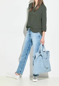 Blouse texturée vert olive, jeans bleu clair et sac fourre-tout bleu clair avec des sangles et une poche avant. Des baskets blanches complètent le look.