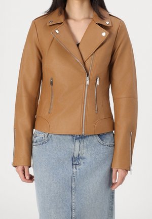 Femme portant une veste de moto en cuir fauve avec des fermetures éclair et des boutons argentés, assortie à une jupe en jean bleu clair.