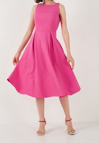 Robe sans manches rose avec un corsage ajusté, une jupe évasée et des plis. Texture lisse, longueur genou, stylée avec des sandales à lanières colorées.