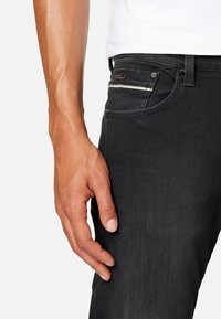 Jeans de mezclilla negra con un ajuste Slim, detalle de cremallera plateada en el bolsillo delantero y una textura suave. Mano descansando ligeramente sobre la tela.