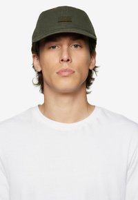 Olive green cap made of cotton with a textured finish, featuring embroidered branding on the front. Worn by a person in a white shirt.