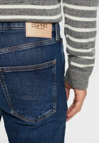 Jean en denim bleu foncé avec un pull côtelé à rayures grises et blanches. Dispose d'une étiquette en cuir beige avec l'inscription "ESPRIT NEW YORK" sur la ceinture.