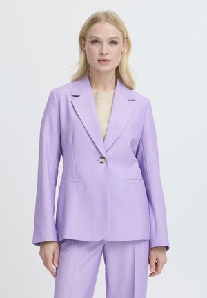 Donna bionda che indossa un blazer lilla su misura con pantaloni coordinati e una maglia beige, in piedi davanti a uno sfondo bianco neutro.