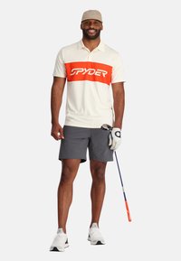 Herren-Polohemd mit kurzen Ärmeln in Creme mit einem auffälligen orangefarbenen horizontalen Streifen, der das "SPYDER"-Logo trägt, kombiniert mit grauen Shorts und weißen Schuhen.