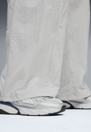Białe trampki ze srebrnymi detalami noszone z luźnymi, jasnoszarymi teksturowanymi spodniami zakrywającymi kostki, stojące na białej powierzchni.