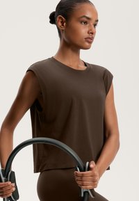 Barna ujjatlan pamut felső kerek kivágással, amelyet mindkét kézben tartott fitneszgyűrű kísér. Simas textúra és laza fazon.