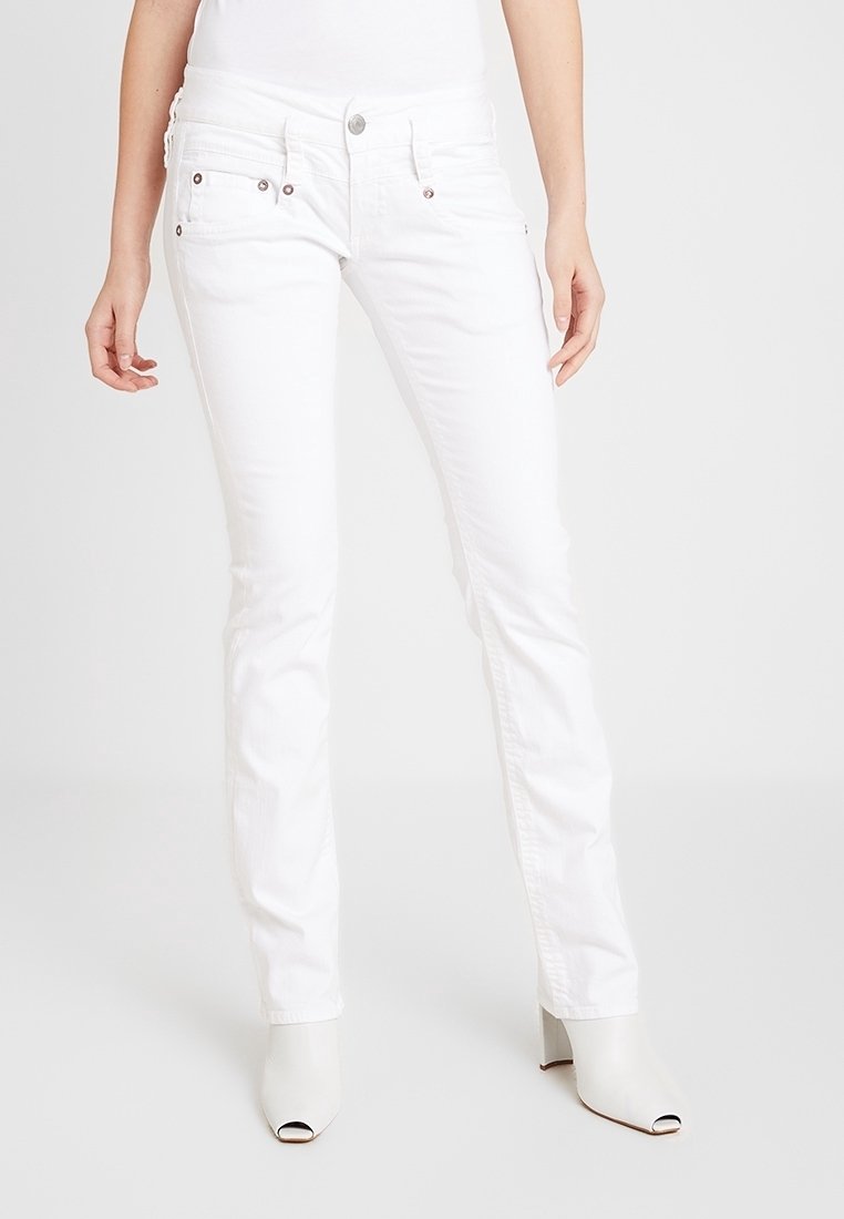 Femme portant un jean blanc slim et des bottines blanches à talons ouverts, debout devant un fond blanc.