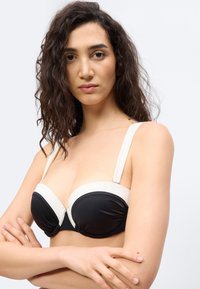 Γυναίκα με σγουρά σκούρα μαλλιά φοράει μαύρο και άσπρο σουτιέν χωρίς τιράντες, σταυρώνει τα χέρια της, κοιτάζοντας απευθείας την κάμερα σε απλό φόντο.