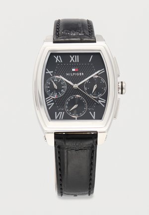 Tommy Hilfiger armbåndsur med sort lærrem, rektangulært sølvfarget urkasse, svart urskive med romertall og tre underskiver som viser 24-timers klokke, dag og dato.