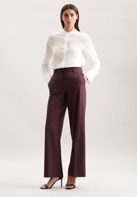 Witte button-up blouse met ribbelstructuur gecombineerd met hooggetailleerde bordeauxrode broek. Model staat in zwarte hakken met handen in de zakken.