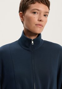 Tamno plava jakna na zatvaranje sa visokim ovratnikom, s glatkom teksturom i minimalnim detaljima šavova. Hardver zatvarača je srebrne boje.