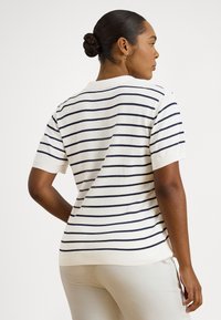 Tumšādainā sieviete ar matiem sasietiem bulciņā valkā baltu īsroku krekli ar plānām melnām horizontālām svītrām un bēšas krāsas bikses, skatoties prom.