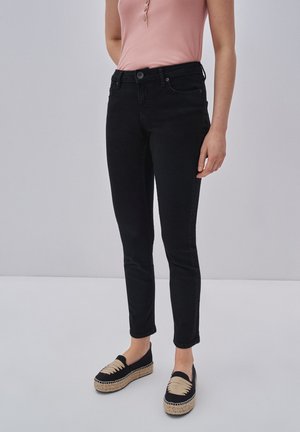 Jeans negros ajustados con una textura suave, talle alto y cierre de botón en la parte delantera, combinados con alpargatas negras con un acento crema.