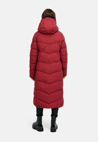 Cappotto lungo imbottito rosso con cappuccio, caratterizzato da un motivo a zigzag e un tessuto testurizzato. Indossato sopra pantaloni neri e stivali neri chunky.