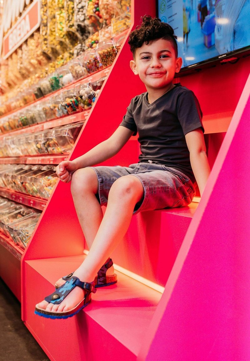 Et barn sidder på en lysende pink geometrisk skulptur, iført en sort t-shirt og grå shorts, med sandaler i blå-og-sorte mønstre.