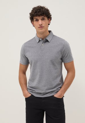 Uomo con capelli ricci che indossa una polo grigia a maniche corte e pantaloni neri, in piedi con le mani nelle tasche davanti a uno sfondo semplice.