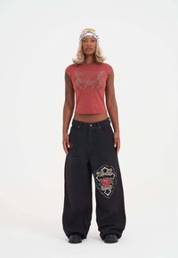 Červený krátký rukávový crop top s květinovou výšivkou, spárovaný s volnými černými denimovými kalhotami, které mají barevnou nášivku a vyšívané detaily.