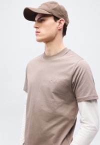 Νεαρός άνδρας που φοράει καφέ γκρι καπέλο μπέιζμπολ και ταιριαστό κοντομάνικο μπλουζάκι πάνω από λευκό μακρυμάνικο, κοιτώντας προς τα αριστερά σε μονόχρωμο φόντο.