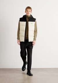 Puffervest med kremfarget kropp og svarte skuldre, kombinert med en tettsittende olivengrønn turtleneck og sorte bukser, brukt med svarte sko.