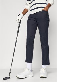 Tengerészkék golfnadrág, szabott fazonban, egy gombbal, fehér sportcipővel és egy golfütőt tartó kesztyűvel párosítva.