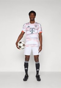 Maglia del Bayern Monaco bianca con motivi marmorizzati rosa e grigi, pantaloncini neri e calzini neri. Il giocatore tiene una palla da calcio. Marchio Adidas.