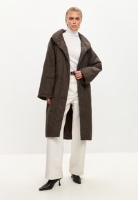 Oversized, mørkebrun quiltet frakke med en bred krave og trykknapper. Båret over en hvid turtleneck, parret med off-white vide bukser og sorte hæle.