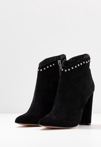Un paio di stivali neri in suede alla caviglia con tacco alto a blocco, cerniere laterali e borchie decorative argentate lungo il bordo superiore.