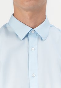 Gros plan d'une personne portant une chemise bleu clair boutonnée avec un col pointu sur fond blanc.