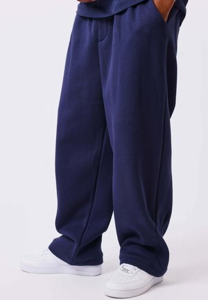 Personne portant un pantalon de survêtement bleu foncé ample et des baskets blanches, debout les mains dans les poches sur un fond clair uni.