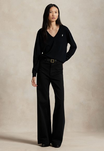 Woman with long dark hair wearing a black V-neck sweater, wide-leg black pants, and black belt, standing against a plain beige background.