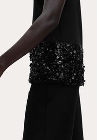 Top negro sin mangas con un ribete negro de lentejuelas en la cintura. Tela suave con un acento texturizado y brillante, vista lateral contra un fondo claro.