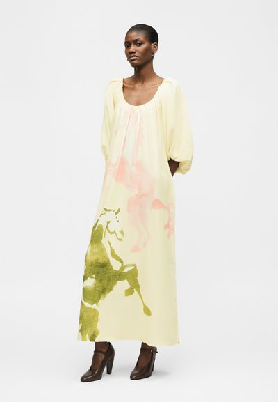 Γυναίκα που φοράει ένα μακρύ ανοιχτό κίτρινο φόρεμα με φουσκωτά μανίκια και σχέδια με άλογα σε πράσινο και ροζ, συνδυασμένο με σκουρό καφέ ψηλοτάκουνα παπούτσια.
