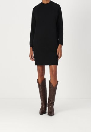 Kobieta ubrana w czarną sukienkę do kolan z długim rękawem, w połączeniu z brązowymi, fakturowanymi kozakami do kolan, stojąca na białym tle.