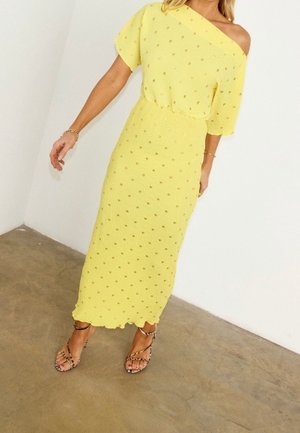 Femme portant une robe jaune, épaules dénudées, longue jusqu'aux chevilles avec de petits motifs à pois et des sandales à brides, debout sur un sol en béton.
