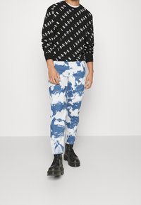 Sudadera negra con texto blanco repetido y pantalones tie-dye azules con patrones de nubes blancas, complementados con botas negras con cordones.