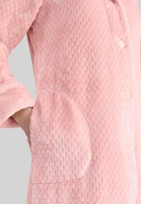 Mano en el bolsillo de una suave chaqueta de forro polar de color rosa claro con botones, mostrando el puño de la manga y el detalle del patrón de la tela.