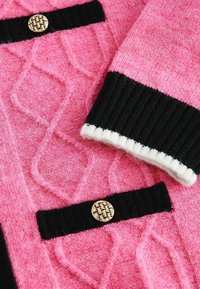 Tissu tricoté rose avec motif losange, poignets côtelés noirs et bordure de poche, chaque poche soulignée par un bouton à motif tissé doré.