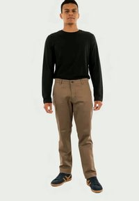 Homme se tenant droit, portant une chemise noire à manches longues, un pantalon marron et des baskets noires avec des accents beiges sur un fond blanc.