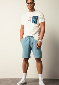 Witte t-shirt met korte mouwen, voorzien van een blauwe grafiek en rode tekst aan de linkerkant, gecombineerd met lichtblauwe shorts met trekkoord en witte sneakers.