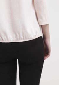 Hellrosa, langärmliges Oberteil mit gerafftem Saum, kombiniert mit schwarzen, körpernahen Hosen. Der Stoff wirkt weich mit glatter Textur und minimalen Details.