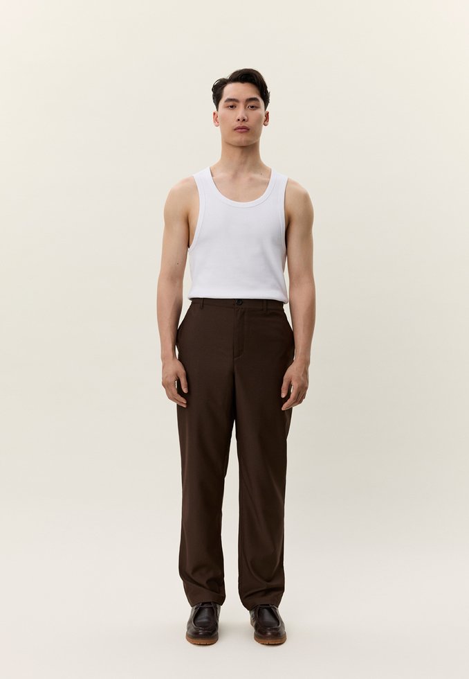 Mladi moški stoji, oblečen v belo majico brez rokavov, temno rjave hlače in temno rjave čevlje, na nevtralnem ozadju.
