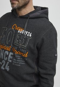 Gray hoodie featuring a drawstring hood, embroidered text in white and orange, and textured fabric. Includes a logo label on the sleeve.